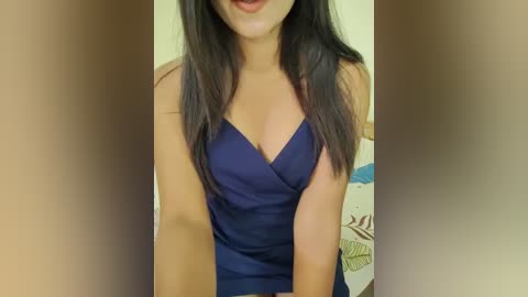Media: A video of a woman with long, straight black hair, wearing a navy blue, sleeveless, V-neck dress, standing indoors against a beige wall with a floral-patterned cushion visible in the background.