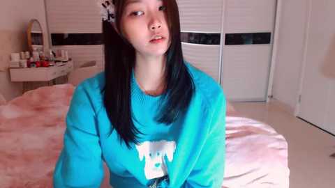 Video of a young Asian woman with long black hair, wearing a blue sweater with a white bear graphic, in a cozy bedroom with a pink blanket, vanity, and white blinds.
