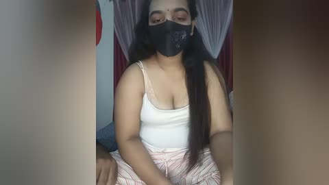 A video of a dark-skinned woman with long black hair, wearing a black face mask, white tank top, and striped pajama pants, sitting against a red and white curtain backdrop.