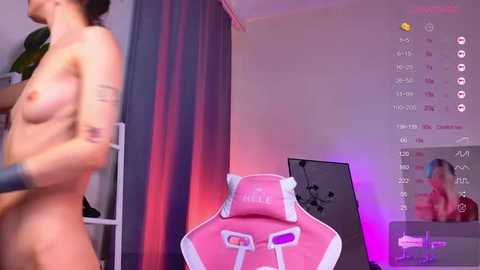 Video of a nude, light-skinned woman with small breasts and tattoos, standing beside a pink gaming chair. Background features a purple-lit room with a calendar and a plant.