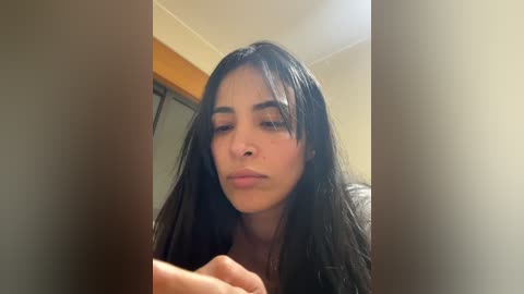 A close-up video of a young woman with long, dark hair and fair skin, wearing a black top, looking contemplative. The background shows a wooden door and beige walls.