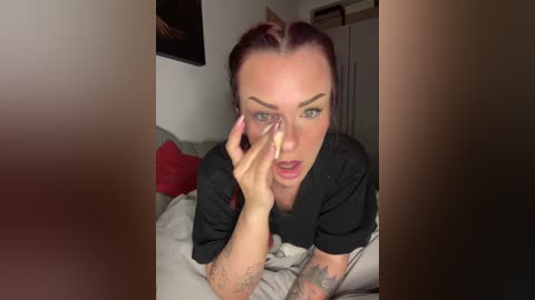 Video of a fair-skinned woman with red hair, wearing a black shirt, lying on a bed, holding her face, showing tattoos on her arms, in a dimly lit room with red pillows and a white wall.