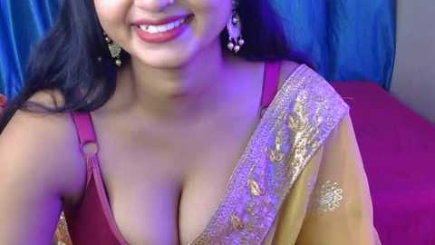 Video of a smiling South Asian woman with long black hair, wearing a gold and red sari adorned with intricate embroidery. She has medium skin tone, wears large gold earrings, and sits against a blue curtain.