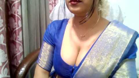 Video of a woman with medium brown skin, wearing a low-cut blue sari with a gold border. She has dark hair pulled back and wears red lipstick. Background includes a patterned curtain and wooden chair.