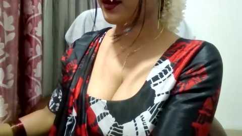 Media: Video of a woman with fair skin and dark hair, wearing a low-cut, black and red patterned blouse with large cleavage. She has a gold necklace and sits against a grey curtain background.