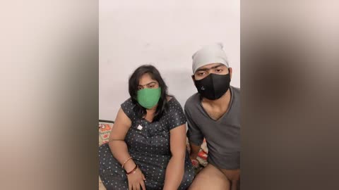 Media: Video of two South Asian individuals wearing face masks, sitting on a patterned couch against a plain white wall; the woman has a green mask, dark hair, and a polka-dot dress, while the man wears a black mask and a gray shirt.
