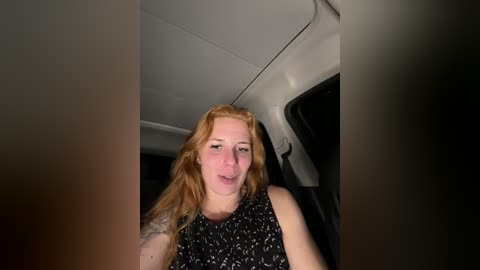 Video of a fair-skinned, red-haired woman with shoulder-length hair, wearing a black floral-patterned sleeveless top, seated in a car with the door open, looking directly at the camera.