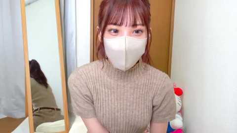 Video of an Asian woman with red hair, wearing a beige ribbed turtleneck sweater, white face mask, and brown belt, standing in front of a wooden door and a mirror, reflecting another person.
