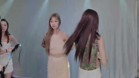 Video of three Asian women with long hair, wearing white dresses, against a soft-focus, light gray backdrop, capturing a moment of playful hair-styling.