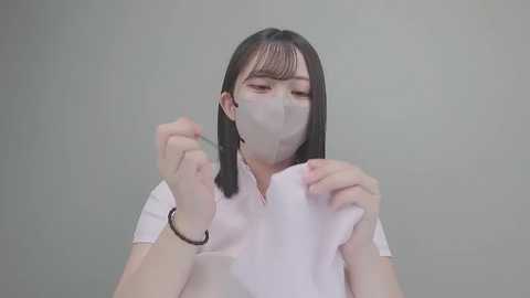 Video of a young Asian woman with straight black hair and fair skin, wearing a white surgical mask and scrub uniform. She holds a white cloth in both hands, against a plain, light gray background.