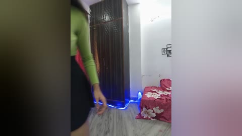 Video of a small, dimly lit room with a person in a green top and black pants walking towards the camera, past a red bed with white floral sheets and a brown partition.