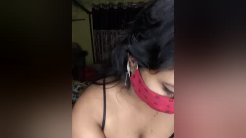 Media: A video of a woman with medium skin tone, wearing a red blindfold and a black spaghetti strap top, her long black hair partially covering her face, in a dimly lit room with a bed and curtains visible in the background.