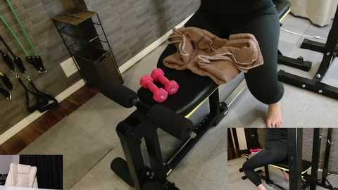 Video of a person in black workout gear sitting on a black exercise bench, wearing a brown towel, with pink dumbbells, in a gym with gray walls and exercise equipment.