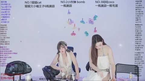 Video of two young women in white dresses, seated on a stage with a backdrop of floating butterflies and Chinese text, surrounded by electronic devices and a black chair.