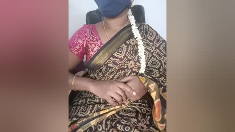 Video of an Indian woman seated, wearing a traditional black sari with gold and red patterns, a pink lace blouse, and a blue mask. She has a white garland around her neck and a gold bracelet on her wrist.