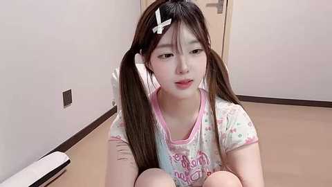 Video of a young Asian woman with long brown hair in pigtails, wearing a pastel-colored T-shirt, sitting on a carpeted floor in a minimalist room with white walls and a closed door.