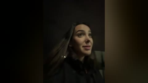 Media: A dimly lit video of a woman with long, straight dark hair, wearing a black cloak, standing in a dark, shadowy hallway. Her face is turned slightly to the right, with a contemplative expression.