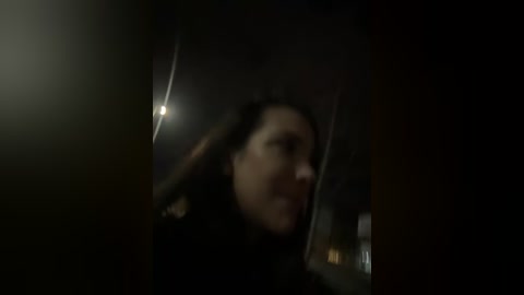 Media: Blurry, low-light video of a woman with long hair, possibly Latina, licking a glass, with a dimly lit room in the background.