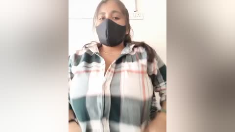 Media: A video of a woman with medium-brown skin and straight black hair, wearing a black face mask, a plaid shirt, and a gold necklace. The background is a plain, light-colored wall with a power outlet.