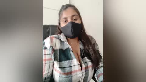 Media: Video of a South Asian woman with long black hair, wearing a black face mask, plaid shirt, seated on a black leather chair against a beige wall.