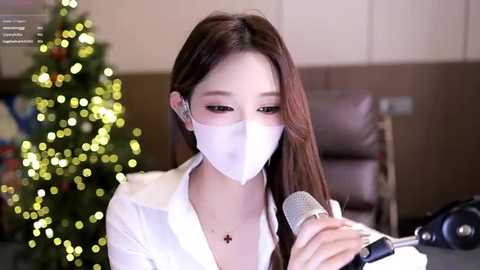 Video of an East Asian woman with long brown hair, wearing a white mask, white shirt, and a microphone, sitting in front of a Christmas tree and brown walls.