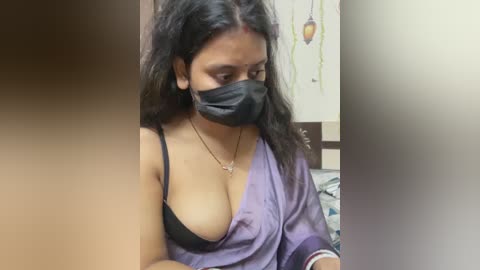 Video of a South Asian woman with long black hair, wearing a black face mask, black bra, and purple satin robe, sitting in a dimly lit room with minimal decor.