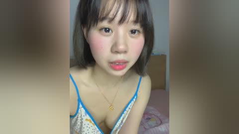 A video of an Asian woman with straight, shoulder-length brown hair, wearing a polka-dotted bra with blue trim. She has light skin, full lips, and a round face, sitting on a pink bed with a floral duvet.