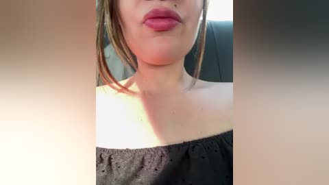 Video of a young woman with fair skin, light brown hair, and full lips, wearing a black off-shoulder top. The background is blurred, showing a car interior.