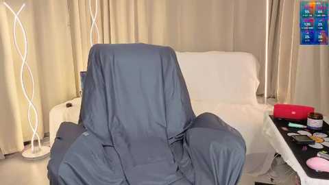 Video of a cozy, well-lit room with a white, covered armchair, beige curtains, a modern lamp, and a colorful calendar on the wall.