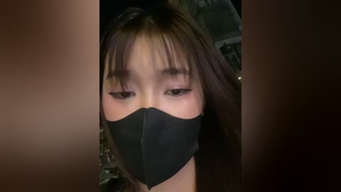 A video of an East Asian woman with long, straight brown hair and bangs, wearing a black face mask, with a blurred background.