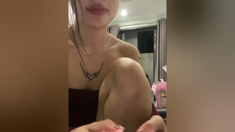 Video of a young Asian woman with light skin, sitting with knees drawn up, wearing a black tattoo necklace and maroon top, in a dimly lit room with a pink toy.