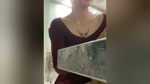 Video of a woman with fair skin, wearing a deep maroon off-shoulder top, holding a mirror, revealing a tribal tattoo on her chest. Background includes a blurred bathroom with a bathtub and shower.