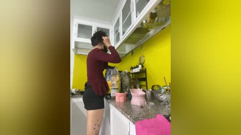 Video of a woman in a kitchen with yellow walls, wearing a maroon top and black shorts, adjusting her hair in front of a granite countertop with pink cups and a bag.