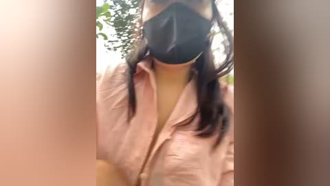 Video of a person wearing a black face mask, light pink button-up shirt, and long dark hair. The background is blurred with greenery, suggesting an outdoor setting. The image appears slightly blurry.