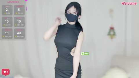 Video of an East Asian woman in a black dress and mask, standing in a modern bedroom with a bed and abstract artwork.