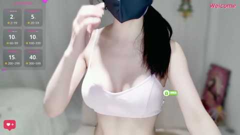 A video of a young, slender woman with fair skin and long black hair wearing a white sports bra and black face mask, taken indoors with a soft-focus background.