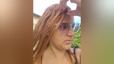 Video of a young woman with light skin and long, wet, reddish-brown hair, wearing clear glasses, holding her hair with one hand, outdoors in a garden setting.