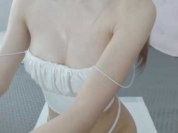 Video of a fair-skinned, slender woman with medium-sized breasts, wearing a white strapless top and white thong, sitting on a grey couch, partially cropped.