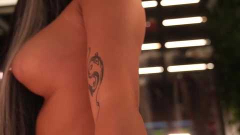 A close-up video of a topless woman with large breasts, showcasing a detailed tattoo of a lion's face on her left arm. The background is blurred, featuring vertical blinds.