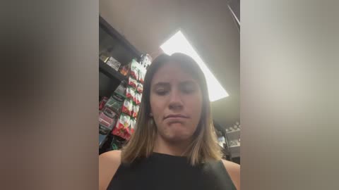 Video of a woman with shoulder-length brown hair, wearing a black tank top, standing in a well-lit, cluttered room. Shelves behind her hold various items, including colorful products.