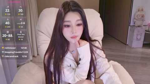 A video of an Asian woman with long, dark hair, wearing a white cardigan, lying on a beige couch, surrounded by a modern, light-toned room with a teddy bear and a clock visible.