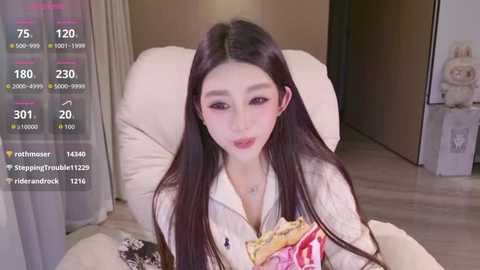 A video of an East Asian woman with long black hair and fair skin, wearing a white robe, holding a box of snacks, sitting on a beige chair.