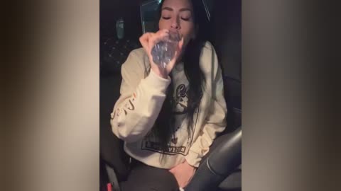 A video of a young woman with long black hair, wearing a beige sweatshirt and black pants, drinking water from a clear plastic bottle while seated in a dark car.