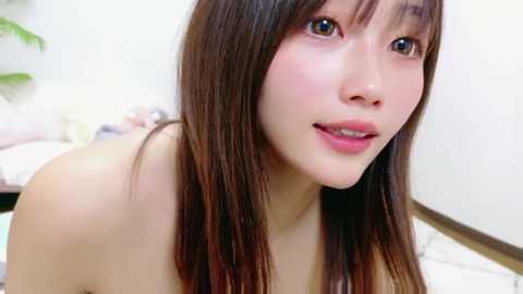 Video of a young Asian woman with long brown hair, fair skin, and large brown eyes, smiling softly, topless in a room with a white wall, blurred background.