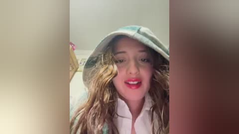 Video of a young woman with long, wavy brown hair, wearing a green and white plaid hooded sweatshirt, smiling with red lipstick, against a blurred beige wall.