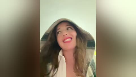 Video of a smiling woman with fair skin, brown hair, wearing a white shirt and a hooded jacket with green and white stripes, looking up, possibly in a room with blurred beige walls.