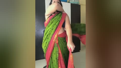 Video of a woman in a green saree with red borders, holding the pallu, standing indoors against a black and white wall. Her dark hair is tied back, and she appears to be Indian.