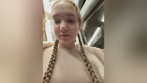 Video of a young woman with blonde braided hair and fair skin, wearing a beige top and a fluffy white jacket, standing in a dimly lit, industrial corridor with metallic pipes and lighting fixtures in the background.