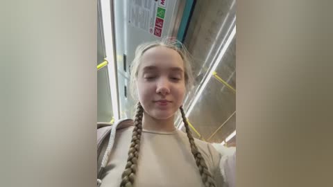A video of a young Caucasian girl with blonde braids, wearing a beige sweater, standing in a subway car. The background shows subway signs and advertisements.