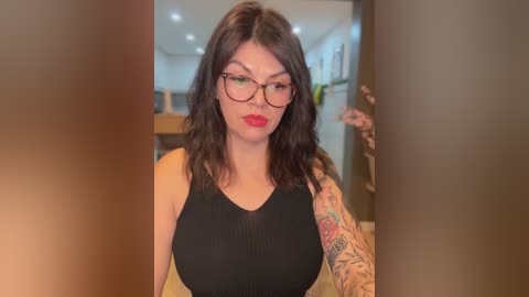 Video of a fair-skinned woman with shoulder-length dark hair, wearing black-framed glasses, red lipstick, and a black sleeveless top. She has a colorful tattoo sleeve and stands in a modern, dimly-lit room with blurred background.
